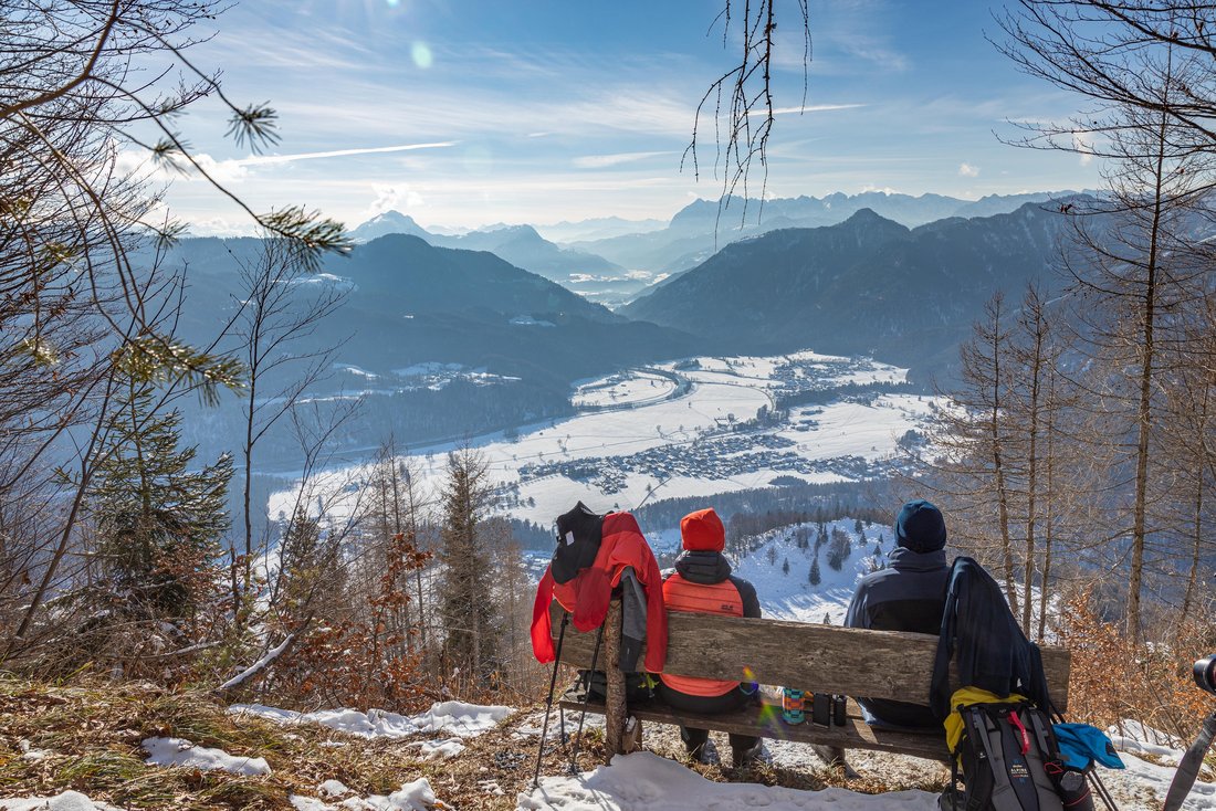 Urlauber sitzen auf einer Bank am Berg und schauen bei Sonnenschein in die verschneite Gemeinde Schleching