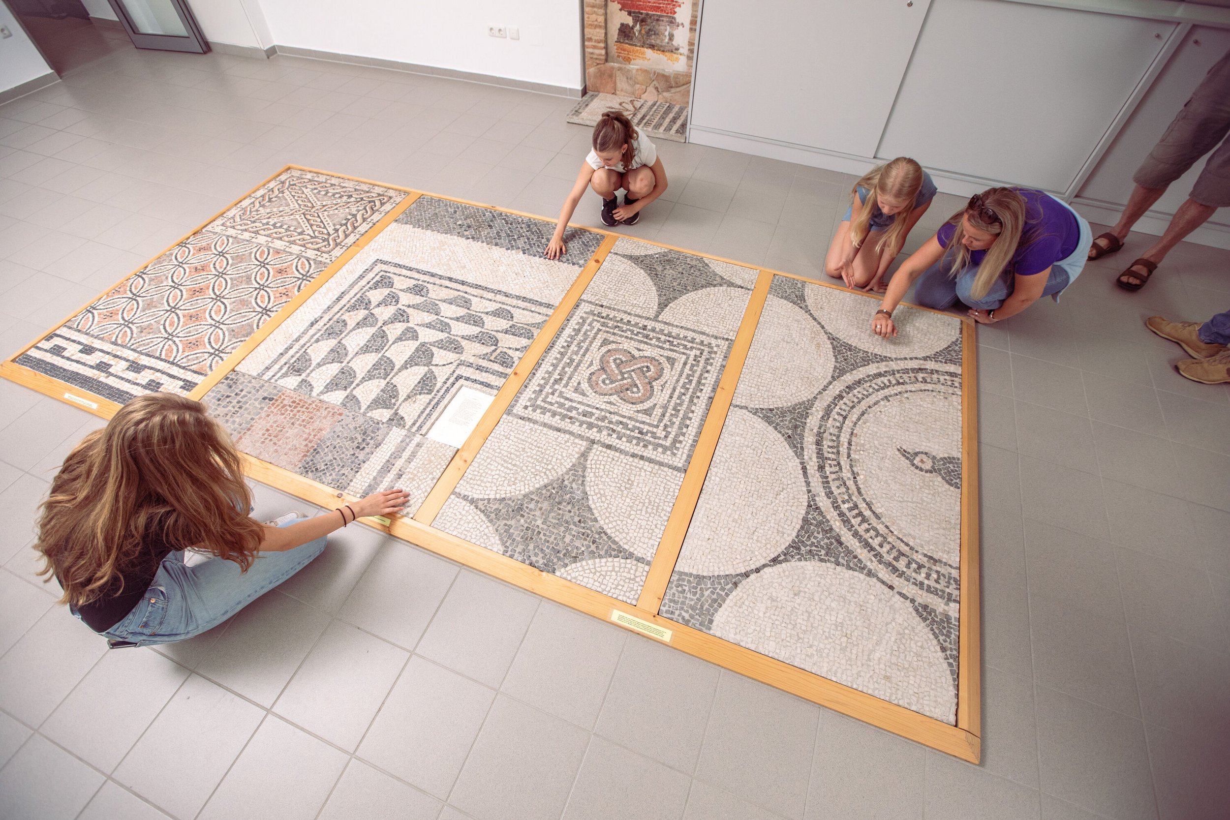 Blick auf einen filigranen Mosaikboden aus der Römerzeit, Frau und drei Mädchen sitzen rund herum
