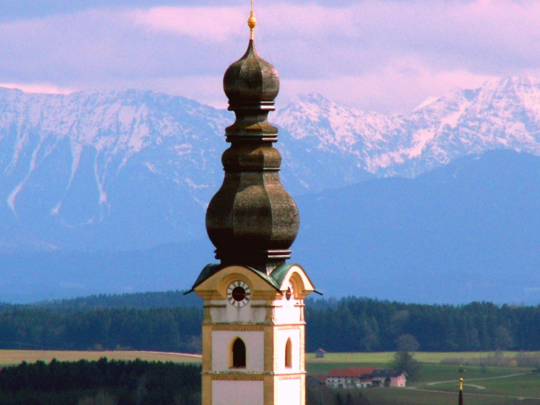 View of the church tower of the Assumption in Schnaitsee with the Bavarian Alps at sunset in the background