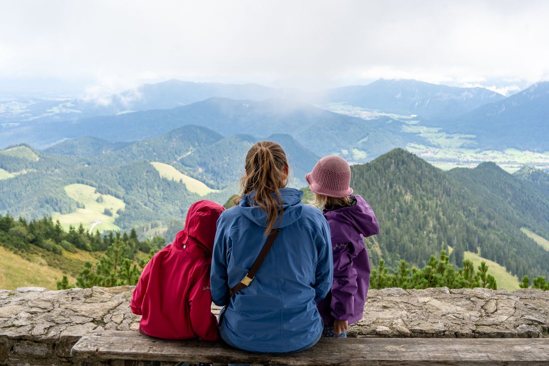 Mutter schaut mit ihren zwei Töchtern von einer Bank bei der Taborkirche am Hochfelln nach Osten ins Tal